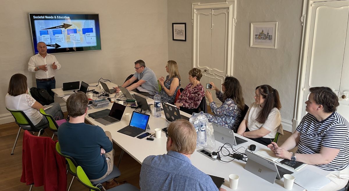 Nine people with laptops in front of them sitting around a table looking towards a man giving a presentation with screen behind him.